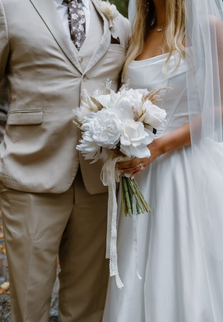 Couple shows off their elopement attire details. A beige men's suit paired with a brown floral tie. The bride wears a vintage gown with white floral bouquet. A beautiful neutral fall color palette for their Great Smoky Mountain elopement.